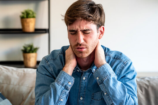 Man in denim shirt holding his neck in pain on a sofa at home, eyes closed and wincing, experiencing neck strain or stress in a cozy living room setting