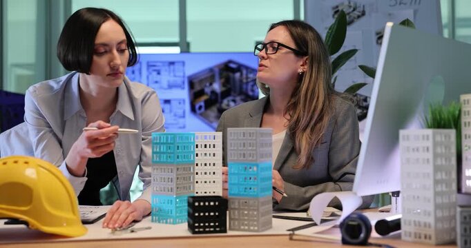 Architectural designers examine detailed models of multi-story buildings. Women discuss elegant urban design solutions in office. Slow motion