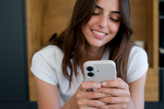 Young woman smiling while using her smartphone at home, typing a message and scrolling social media, enjoying casual communication and digital connection in a relaxed indoor setting.