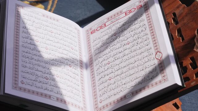 Close up static shot of the Holy Quran open on a carved wooden rehal stand. Bright natural daylight casts dramatic shadows across the Arabic text in a peaceful indoor prayer setting.