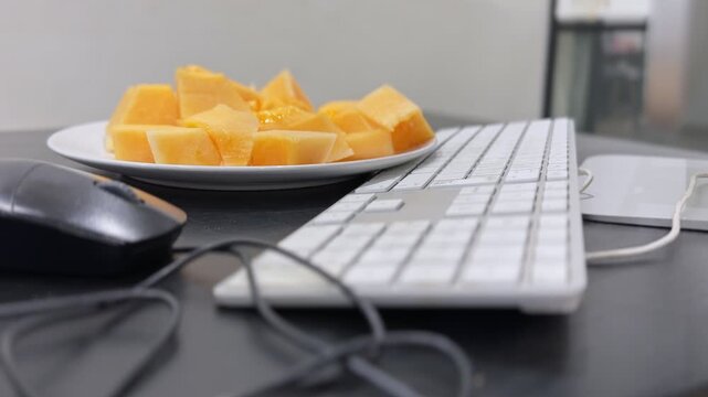 Close up static shot of fresh sliced cantaloupe melon on a white plate resting on a black office desk. A wired mouse and white keyboard sit in the blurred foreground in soft indoor lighting.
