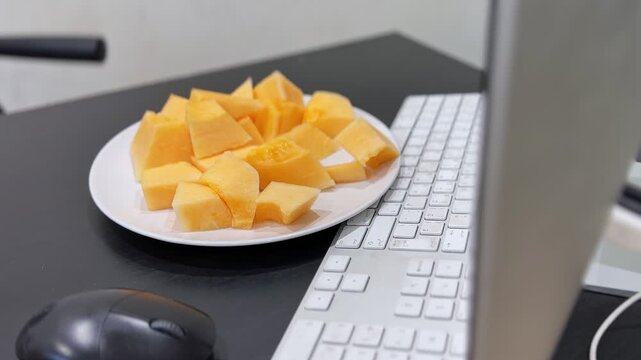 Close-up of a white plate filled with fresh sliced cantaloupe resting on a black modern computer desk. A white keyboard and mouse sit nearby in soft indoor lighting, highlighting a healthy work snack.