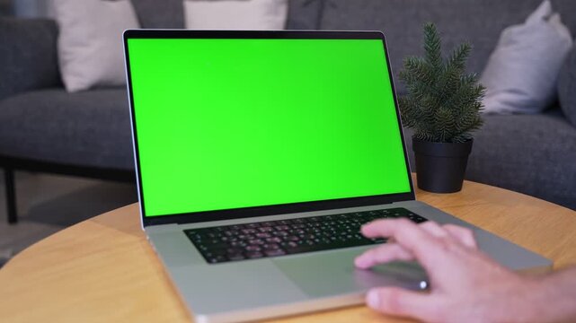 Close up shot of a hand using a laptop trackpad with a blank green screen display. The computer rests on a wooden coffee table in a modern living room with a blurred grey sofa in the background.