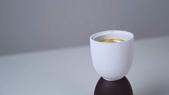 Close up shot of a minimalist white ceramic incense burner emitting a faint trail of smoke. The textured mabkhara sits on a clean white table against a soft grey studio background indoors.