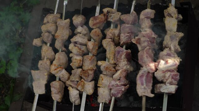 Pork shish kebab on metal skewers appetizingly roasting on a mangal, top view. The process of cooking marinated meat over hot wood charcoals with white smoke.