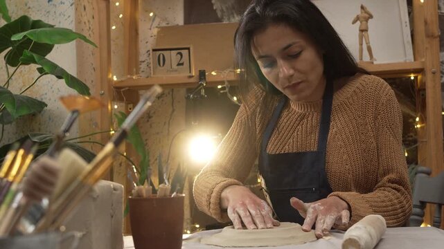 A female potter sits in an authentic workshop, sculpting a clay plate with her hands. The concept is hobby and leisure.