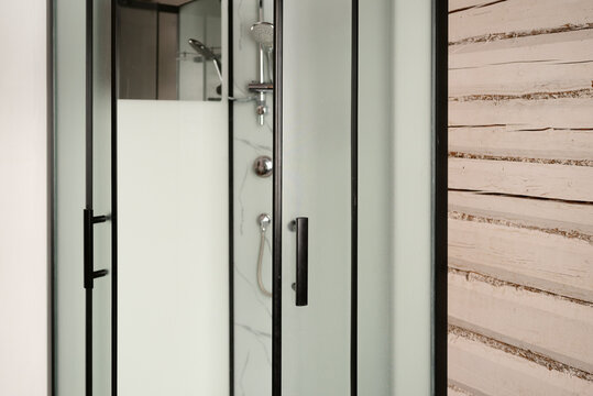 Modern corner shower enclosure with frosted glass doors and metal plumbing fixtures installed against a rustic wooden log wall. Illustrating bathroom interior design and daily hygiene.