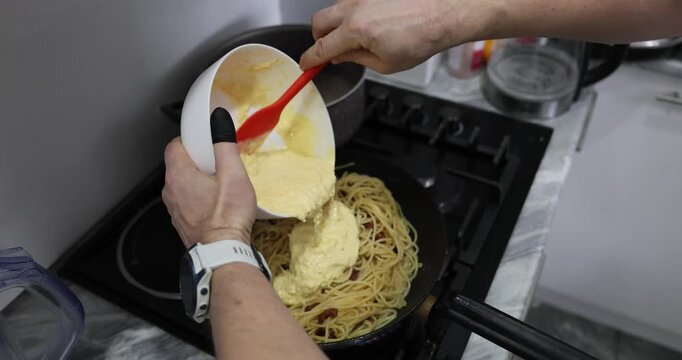 Chef pours sauce over tender spaghetti and golden bacon pieces in frying pan. Experienced cook prepares delicious carbonara dish. Slow motion