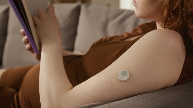 Close up on arm of pregnant Caucasian young adult woman wearing continuous glucose monitor while reading book on sofa. Clip perfect for gestational diabetes and prenatal healthcare marketing