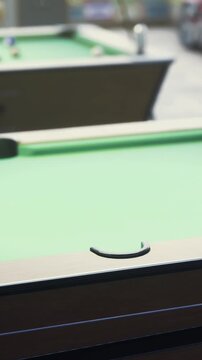 Two pool tables are set up in a recreation center. Players are gathered around, focusing on their shots. The activity takes place in the afternoon, under bright lighting.