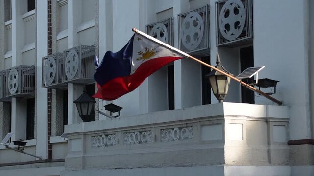 Philippine flag moving with the wind in a sunny afternoon. Red, blue, white, and yellow. Three stars and a sun. Building with white paint.