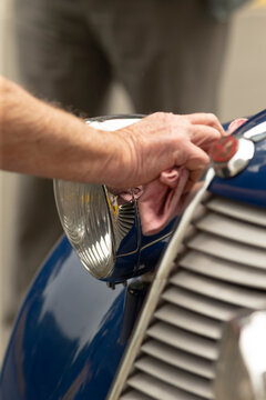 Close view of a person&rsquo;s hands polishing the chrome headlight and adjacent grille of a glossy blue vintage car with a soft cloth. The scene shows metal