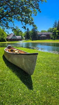 Canoe resting on green grassy shore near park pond and buildings