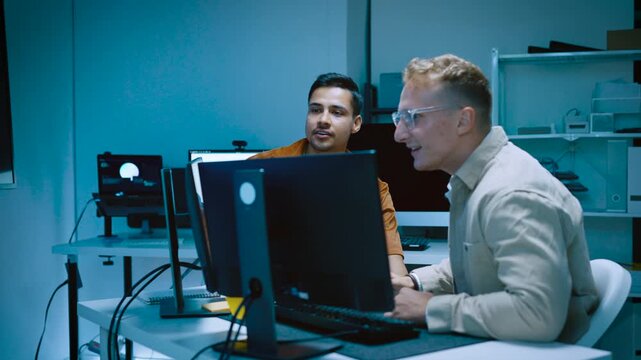 Two young male IT software developers high five celebrating successful coding project completion while working with computers in modern tech office workspace late at night