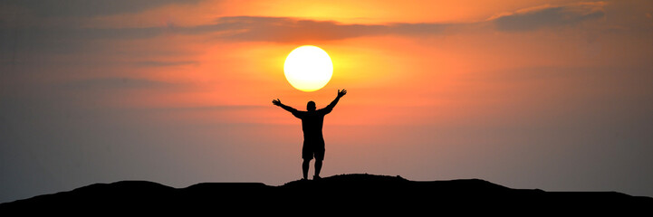A silhouette of a person with arms raised to the sky, embracing the sunset. The person stands atop a hill, with the sun setting behind them, casting a warm, orange glow across the clouds. ©  2026stockx