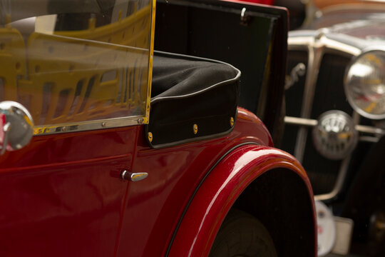 Detailed close-up of a red vintage automobile showing a glossy curved front fender, black leather fold-down seat with brass fasteners, yellow-tinted windshield