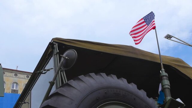 Bottom up view of waving American flag mounted on vintage army truck, evoking patriotism, military history and remembrance against a partly cloudy sky