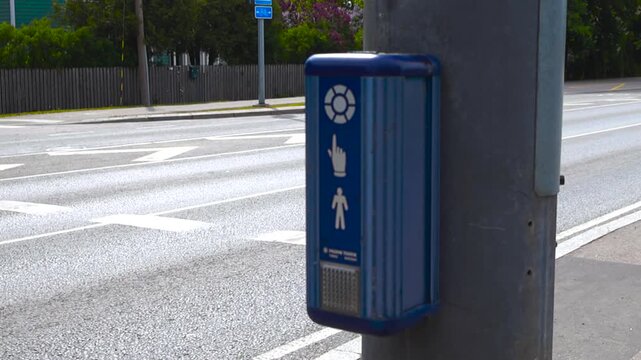 Close up of a blue pedestrian traffic light button at a city intersection. With the yellow wait light blinking to signal that the request has been registered and a crosswalk visible in the background