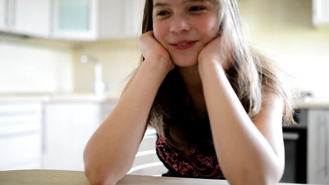 A smiling girl sits at the table on a bright kitchen.