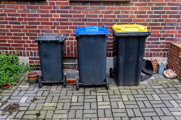 Obraz premium Row of three different sized garbage and recycling plastic containers standing outside on a cobblestone pavement next to a red brick wall.