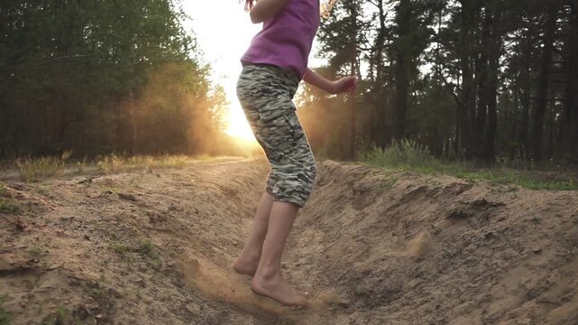 Little girl in camouflage pants jumps through sandy moat in pine forest. Slow motion. Evening time. Sunset.