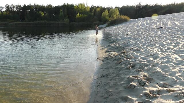 Beautiful little girl with long hair runs along water near sandy shore of small lake.