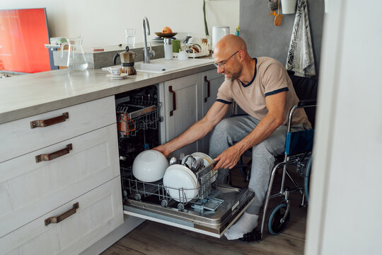 happy smiling man in wheelchair loading or unloading dishwasher in bright modern kitchen side view representing independent living adaptive lifestyle household chores accessibility inclusion