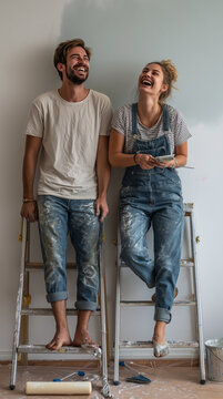 Two adults laughing on ladders during interior painting, paint-splattered clothes and tools on drop cloth