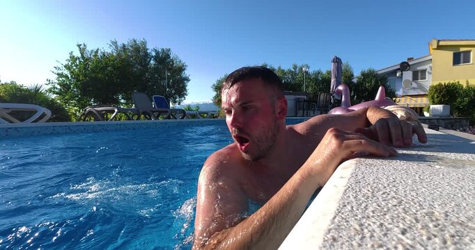 Man swims across the pool and reaches the edge, breathing heavily and recovering after physical effort. Authentic summer scene featuring exercise, endurance, and relaxation by the water.