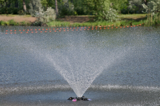 Decorative floating water fountain aerator splashing in a pond on a summer day. Fresh droplets spray over the water surface against a natural green shoreline background. Photo