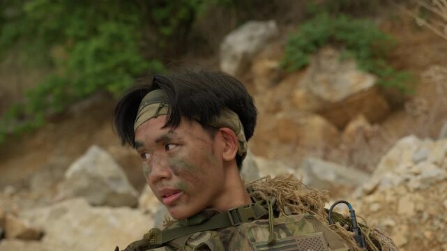 A focused young soldier in tactical gear with camouflage paint conducts surveillance during a military training mission in a rugged, rocky terrain.