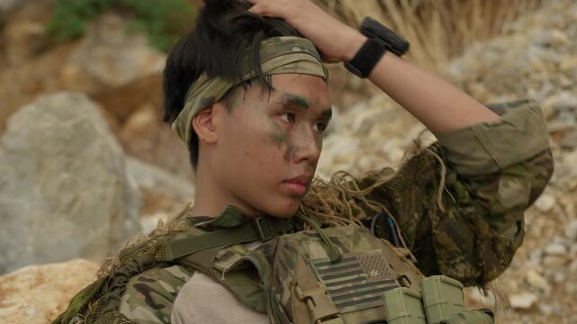 A young Asian soldier with face paint in full tactical gear remains vigilant while on a reconnaissance mission in a rugged, hostile outdoor environment.