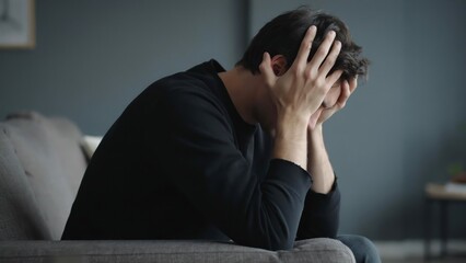 Obraz premium Man holding head in hands sitting on couch in dimly lit room expressing stress and anxiety