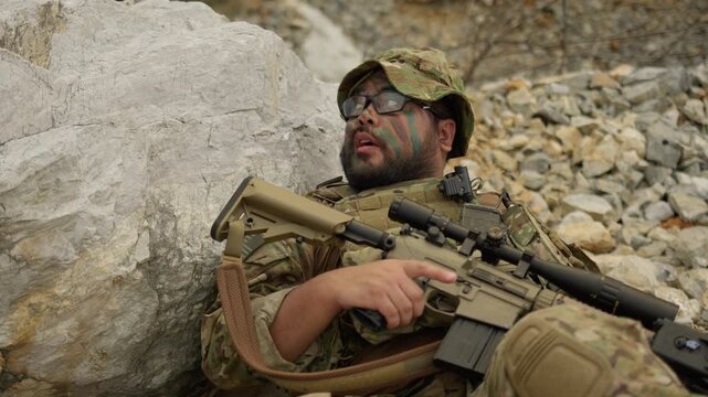 An exhausted special forces soldier rests against a rock in a hostile environment, holding his rifle and showing the heavy toll of a military mission.