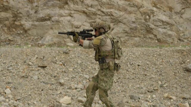 A special forces soldier in full tactical gear patrols through a rocky quarry, aiming his assault rifle during a military training operation or combat mission.