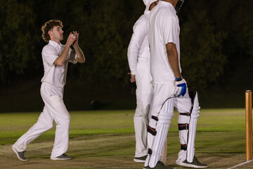 Obraz premium Male cricketers in white kit appealing near bat and stumps on pitch under floodlights, copy space