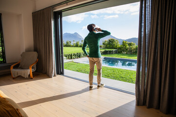 Naklejka premium Man is standing by sliding glass door on wood floor, talking on smartphone, pool visible
