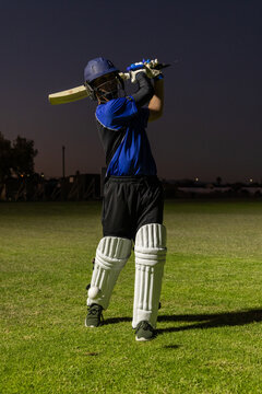 Cricket batsman is holding bat in follow-through on grassy pitch at dusk with ball near pad