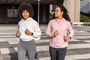 Two female friends jogging across tiled plaza, passing columns and wearing fitness watches © wavebreak3