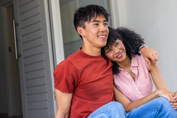 Obraz premium Diverse couple sitting on home's entrance steps by doorway, leaning together and wearing smartwatch