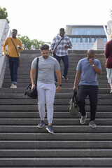 Fototapeta premium Diverse male group descending stone stairway in city, carrying duffel bags and takeaway cups