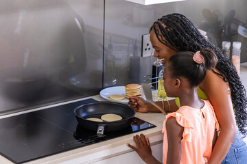 African American mom and daughter cooking pancakes at stove using spatula and pan, copy space © wavebreak3
