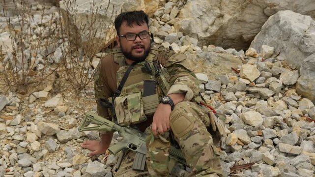 A man in tactical gear takes a break from a military simulation, resting with his rifle among the rocks of a rugged, outdoor training environment.