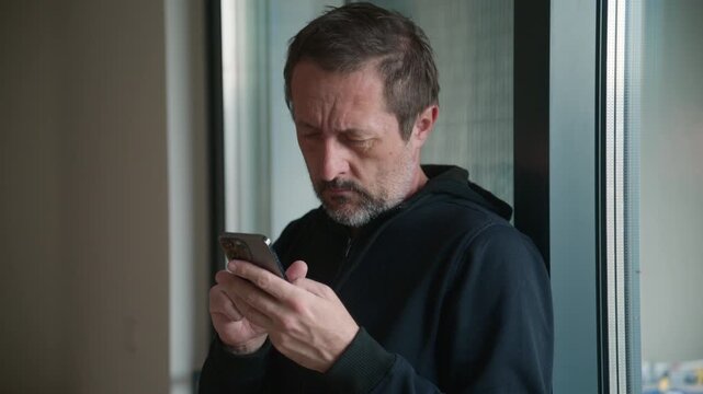 Mature bearded man leaning on apartment window while typing message on smartphone, casual everyday communication at home, 4K