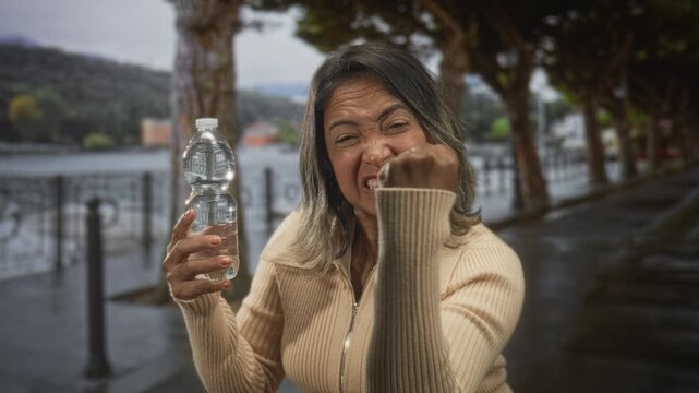 Woman middle age hispanic holding a plastic water bottle, squinting and making a fist with hand along a waterfront street lined with trees and railing; defiance.
