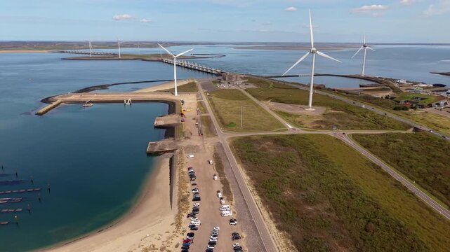 Aerial view of a coastal dam with wind turbines, a sandy beach, parked vehicles, and water channels in a delta landscape under clear daylight.
