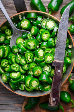 Metal pot filled with sliced jalapeno peppers and knife on wooden surface top view. Fresh green chili rounds with seeds rustic kitchen preparation