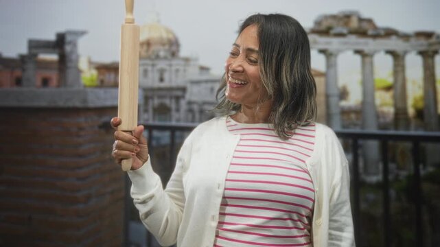 Woman holds rolling pin with hand on balcony overlooking ancient ruins and stone columns, smiling at the tool; home baking joy.