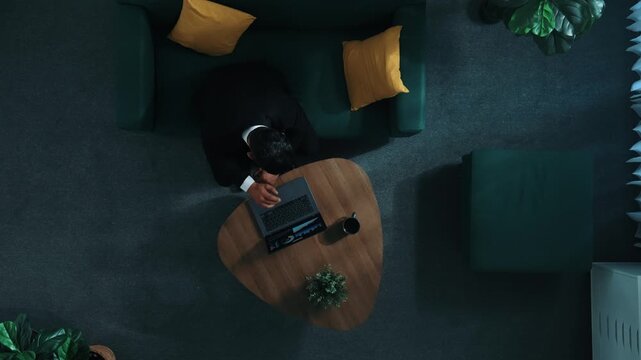 Top down aerial view of stressful boss look at decrease sales from laptop and lies on sofa. Tired manager working laptop while feeling stress. Skilled businessman having headache at work. Directorate.