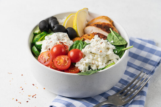 Healthy high protein Mediterranean chicken bowl with rice, cucumber, cherry tomatoes, feta cheese, olives, spinach and creamy yogurt sauce on light background with copy space